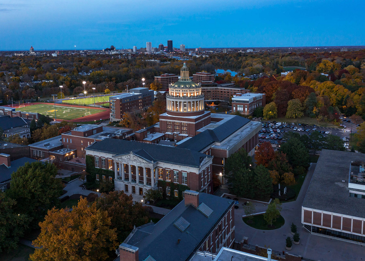 nighttime aerial view of Rush Rhees library with city skyline in distance.